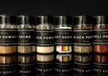Image caption: Studio Barnes. Spices from A Spectrum of Blackness: The Search for Sedimentation in Miami. 2020. Plastic, spices, paper, and adhesive Image description: Photo of six bottles of spices against a black background and a reflective black surface. Each bottle has a black label with white text. The labels read from left to right: Trinidadian/ Curry Powder/ to win the curry duck competition; Hatian/ Thyme/ Core Spice; Bahamian/ Onion Powder/ Put this on your conch fritters; African-American/ Hot Sauce/ Yes, Hot Sauce is a spice; Jamacain/ Black Pepper/ Core Spice; Hondorun/ Chili Powder/ in your Montucas, it’s like tamale.